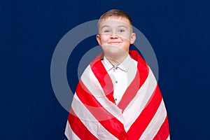 Happy young caucasian american boy kid child schoolboy wraps himself in a U.S. flag and look at camera