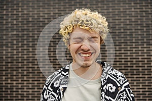 Happy young caucasian blond man with closed eyes smiles with teeth against wall background.