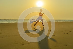 A happy young caucasian blonde woman in sunglasses and a fluttering white dress runs down the beach at sunset