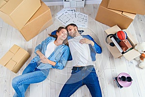 Happy young caucasian couple lying down on the wooden floor while having a break while moving to new house. Top view.