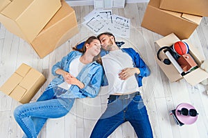 Happy young caucasian couple lying down on the wooden floor while having a break while moving to new house. Top view.