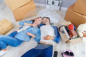 Happy young caucasian couple lying down on the wooden floor while having a break while moving to new house. Top view.