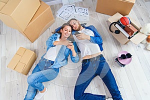 Happy young caucasian couple lying down on the wooden floor and messaging at smart phones during having break while