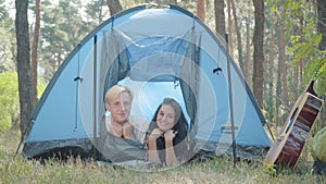 Happy young Caucasian couple lying in tent in forest and smiling at camera. Portrait of relaxed handsome blond man and