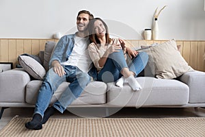 Happy young Caucasian couple relax on cozy sofa at home hugging look in distance dreaming together