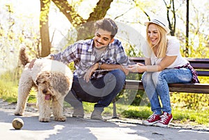 Happy young caucasian couple sitting on a park bench, with a dog and having fun on a sunny day