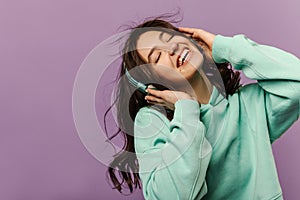 Happy young caucasian girl with eyes closed, holding headphones on head against purple background.