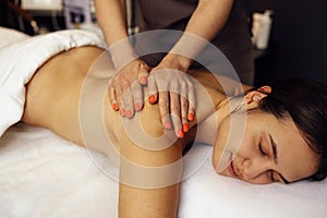 Happy young caucasian girl getting back massage in spa salon. Neat hands of female masseuse with orange manicure