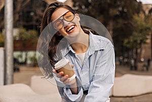 Happy young caucasian girl laughing looking at camera, holding glass of coffee walking in street.