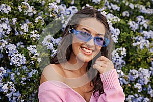 Happy young caucasian girl smiles teeth looking at camera on background of blue flowers on bush.