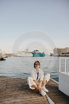 Happy young caucasian girl in sunglasses relaxing on weekend sitting pier by sea.