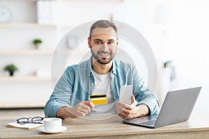 Happy young Caucasian man shopping online, using smartphone, laptop and credit card at home office