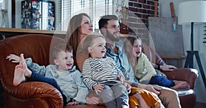 Happy young Caucasian mother, father and three children smile watching TV at home together on the couch slow motion.