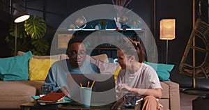 Happy young Caucasian woman and black man, student friends work together using laptop talking, sitting on floor at home.