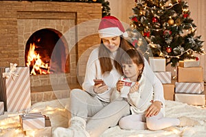 Happy young Caucasian woman with her little daughter girl having fun on floor, using mobile phone, celebrating Christmas at home,
