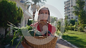 Happy young Caucasian woman is holding a basket with fresh biologic just harvested vegetables and smiling in camera on