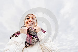 Happy young Caucasian woman looking at the camera wearing white woolen cap, knitted braids sweater with colored scarf in winter