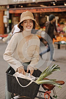 Happy young caucasian woman looks at camera, puts her purchases in bicycle basket.