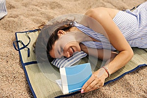 Happy young caucasian woman lying on underlay and reading book on beach in sunny weather.