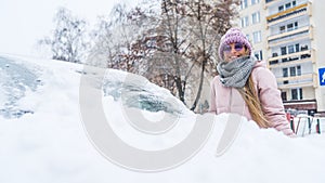 Happy young caucasian woman in pink jacked cleaning irst snow from her car