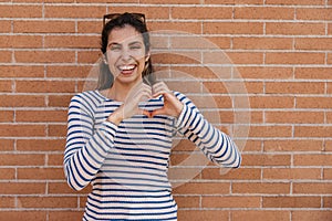Happy young caucasian woman smiles teeth, makes heart gesture with hands standing near brick wall.
