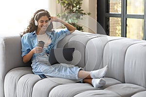 Happy young Caucasian woman using laptop and headphones, having online video call, drinking coffee at home