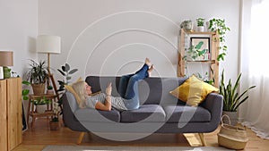 Happy young caucasian woman using mobile phone while lying a couch at home.