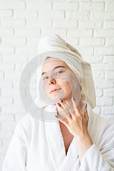 Happy young caucasian woman in white bath towel applying face cream at home