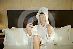 Happy young Caucasian woman in white bathrobe and wrapped head in bath towel lying on the bed in hotel bedchamber and smiles