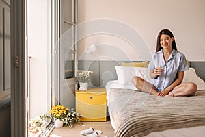 Happy young caucasian woman with wide smile looks at camera and sits on bed near window.