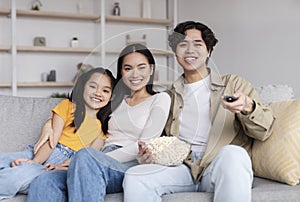 Happy young chinese wife, husband and teenage girl watching tv, eating popcorn, sitting on sofa in living room interior