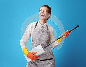 Happy young cleaning woman in apron on blue using mop as guitar