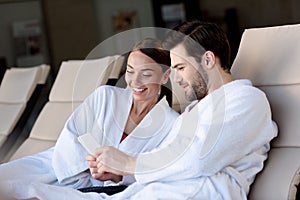 happy young couple in bathrobes using smartphone together while resting