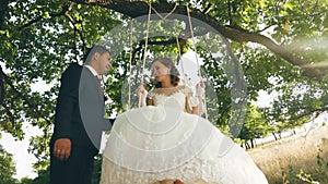 Happy young couple bride and groom in wedding dress swinging on swing in park. swings on branch of an oak in summer
