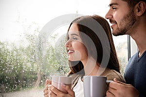 Happy young couple with cups near window on rainy day