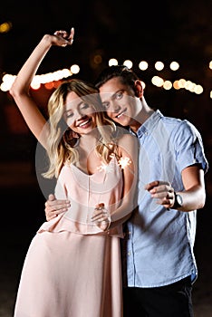 happy young couple dancing and holding party sparkles on river beach