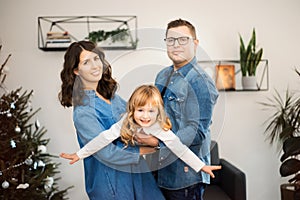 Happy young couple with daughter celebrating Christmas together at home near Christmas tree. Man and woman hold flying girl.