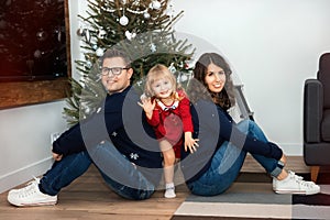 Happy young couple with daughter celebrating Christmas together at home near Christmas tree. Man and woman sitting and girl