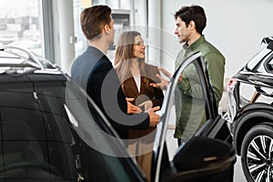 Happy young couple discussing purchase of new car with auto salesman at vehicle dealership
