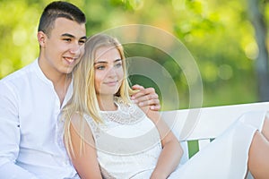 Happy young couple dressed in white looking the same direction on a park bench smiling. Boy holding the girl around her shoulder