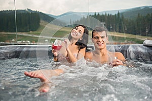 Happy young couple enjoying a bath in Jacuzzi while drinking cocktail outdoors on romantic vacation