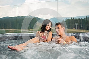 Happy young couple enjoying a bath in Jacuzzi while drinking cocktail outdoors on romantic vacation