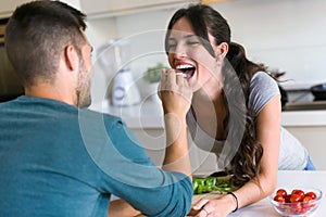 Happy young couple enjoying lunch while handsome man is feeding with yogurt his wife in the kitchen at home.