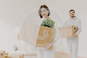 Happy young couple enter own modern house, buy real estate, carry cardboard boxes with indoor plant and other personal stuff, buy