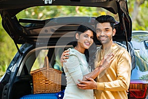 Happy young couple in front of car with travel bags and picnic basket standing by embracing each other while looking at