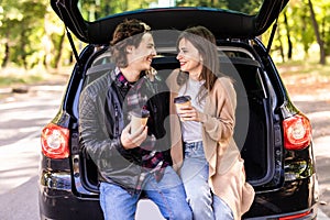 Happy young couple having a coffee break during road trip in countryside. Man and woman sitting in car trunk and having coffee