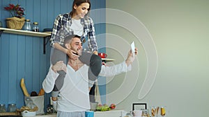 Happy young couple having fun and talking online video call in the kitchen at home. Girl is sitting on boyfriend`s neck