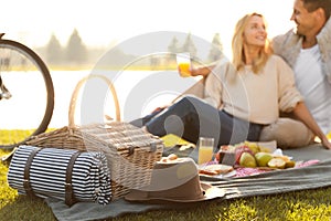 Happy young couple having picnic near lake, focus on basket with blanket and hat