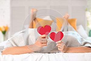 Happy young couple hiding under blanket while holding red hearts on bed at home