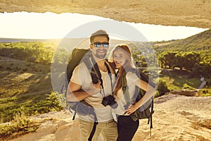 Happy young couple of hikers standing in mountains cave at sunset. Tourists mountaineering on summer vacation